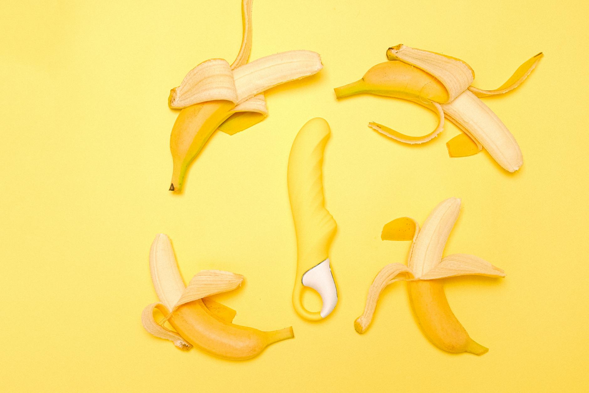 Kreative Flach-Ansicht eines gelben Silikonvibrators umgeben von geschälten Bananen auf gelbem Hintergrund, das Vergnügen symbolisierend.