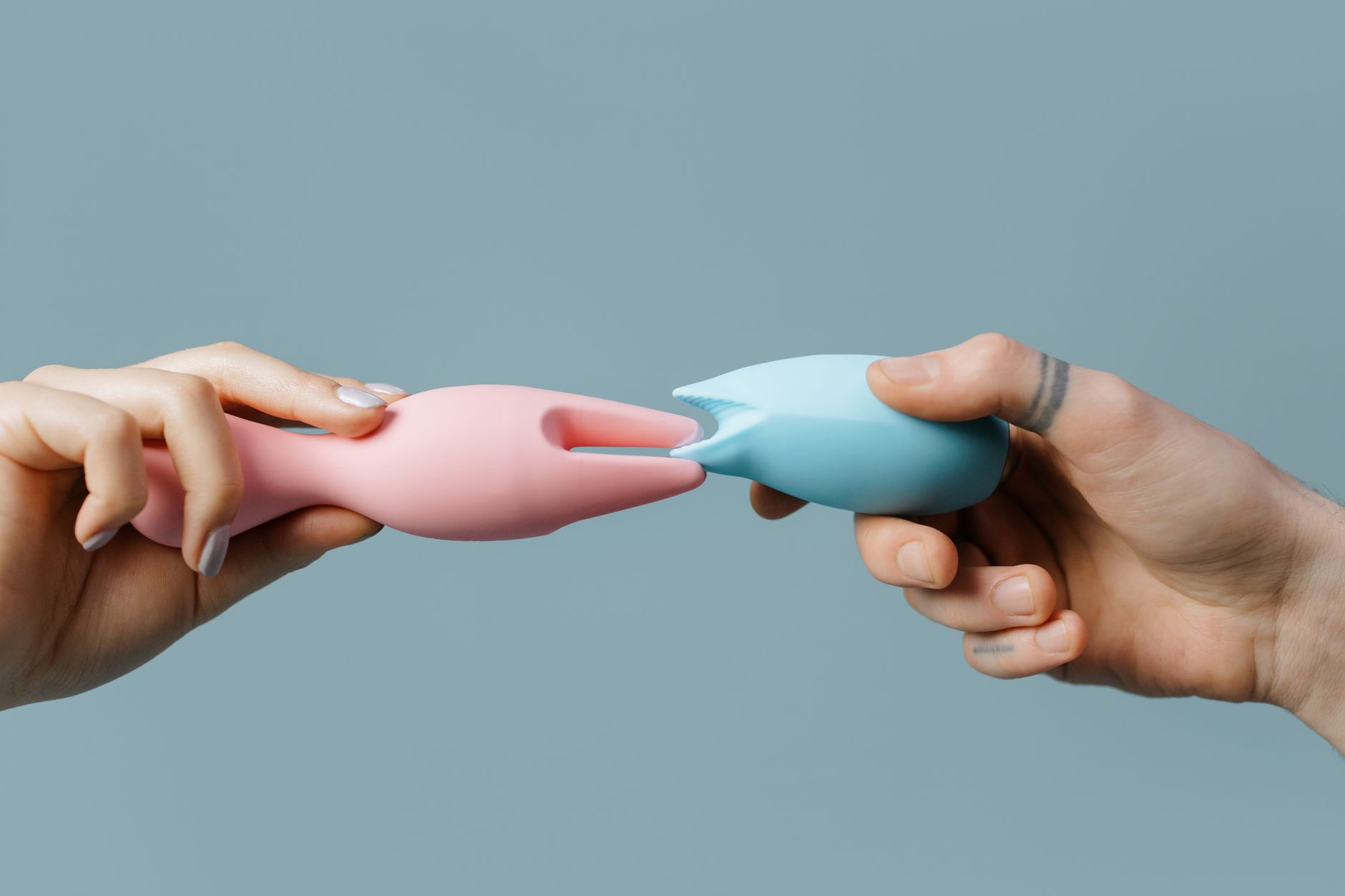 Zwei Hände halten rosa und blaue Silikonvibratoren gegen einen Pastellhintergrund