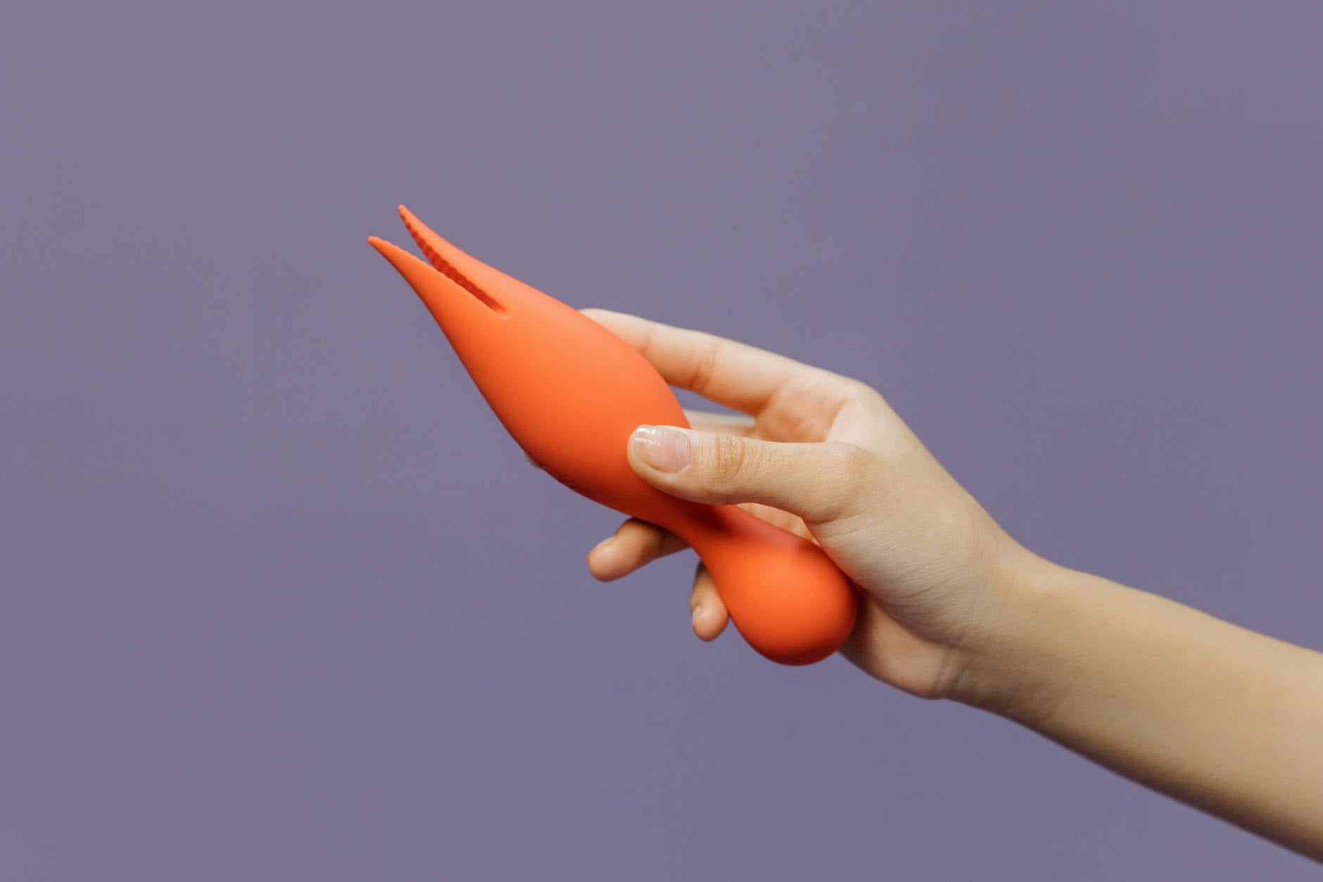 Eine Hand hält einen orangefarbenen Vibrator gegen einen minimalistischen violetten Hintergrund und zeigt moderne Sinnlichkeit.