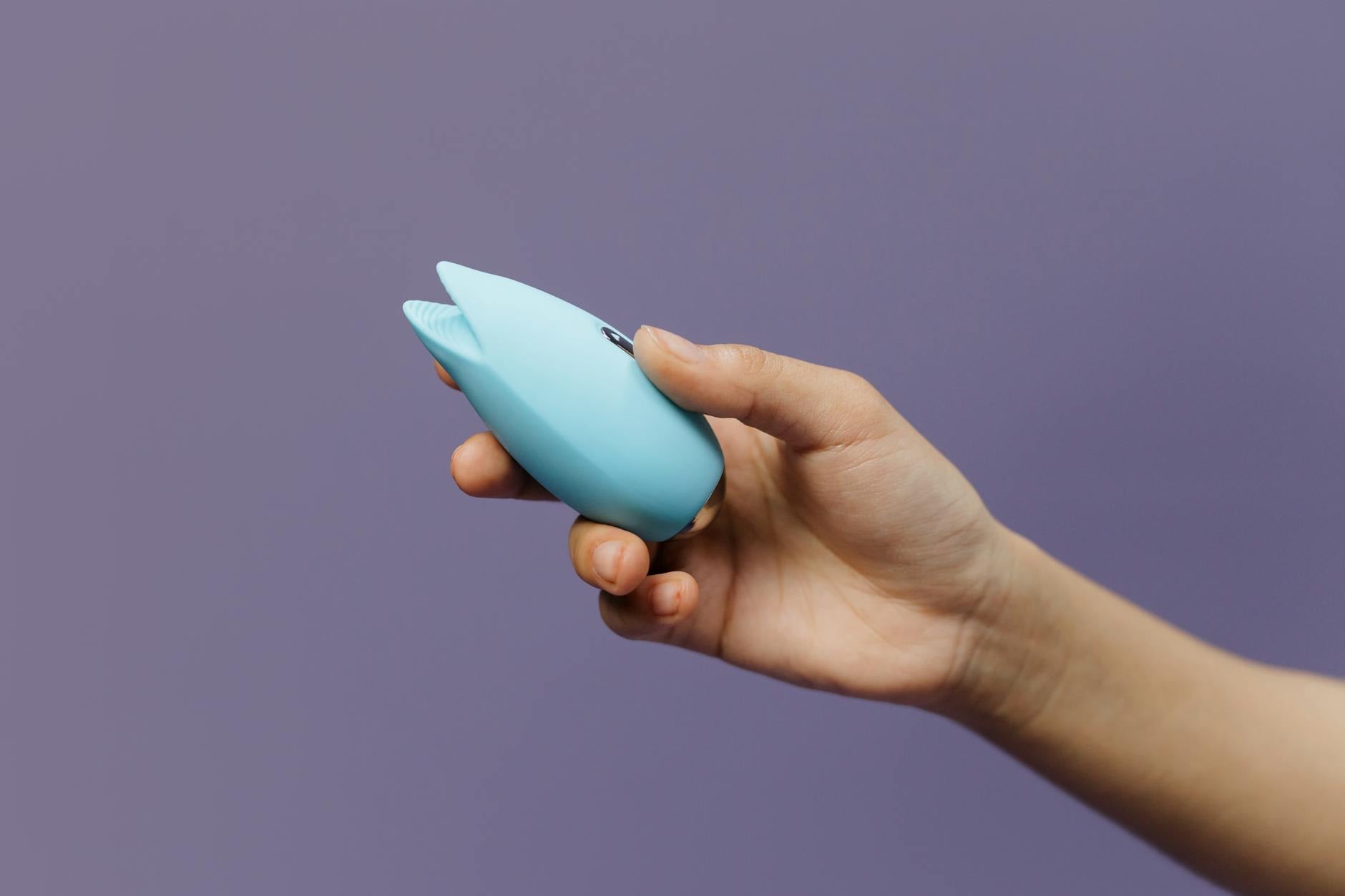 Ein Vibrator wird in einer Hand gehalten gegen einen violetten Hintergrund, der Selbstliebe und Sexualität ausdrückt
