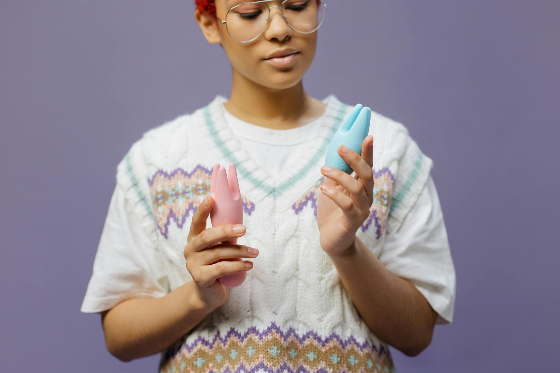 Frau mit Brille hält einen blauen und einen rosa Silikonvibrator nachdenklich in den Händen.